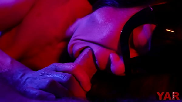 Eine maskierte Frau lutscht einen großen Schwanz und gibt im Mund einen sanften Blowjob im Neonlicht zur Musik